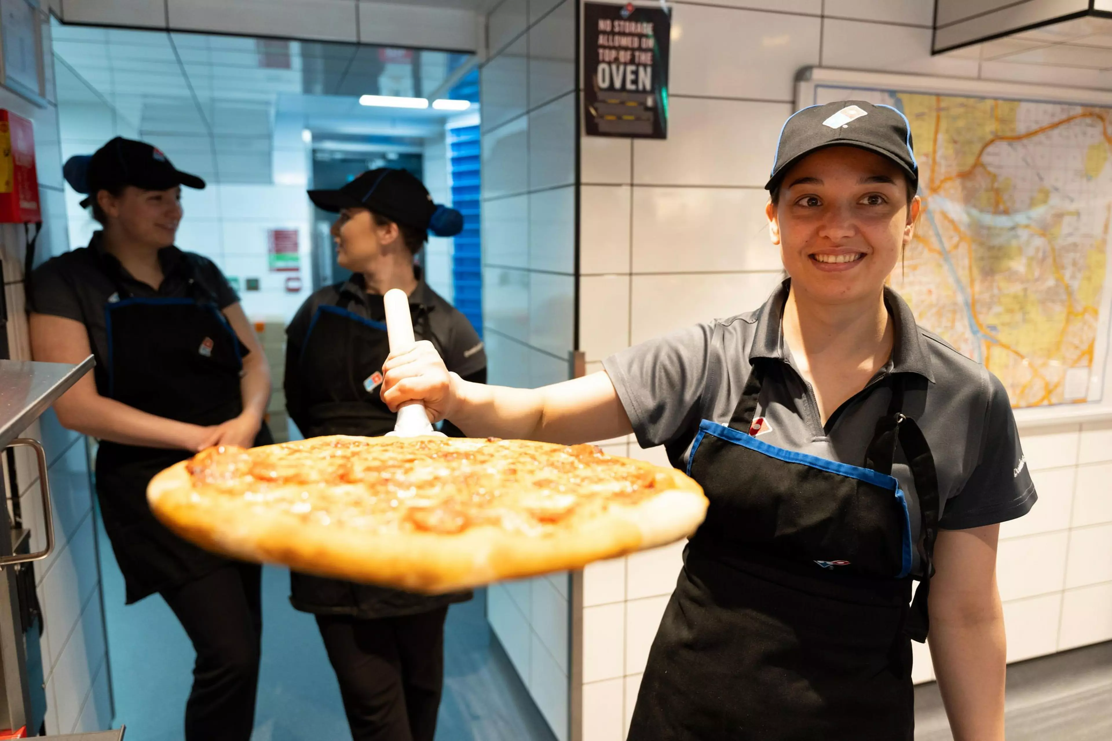 3 Domino's workers stand in a store, two stand in the background talking whilst one in the foreground holding a pizza.