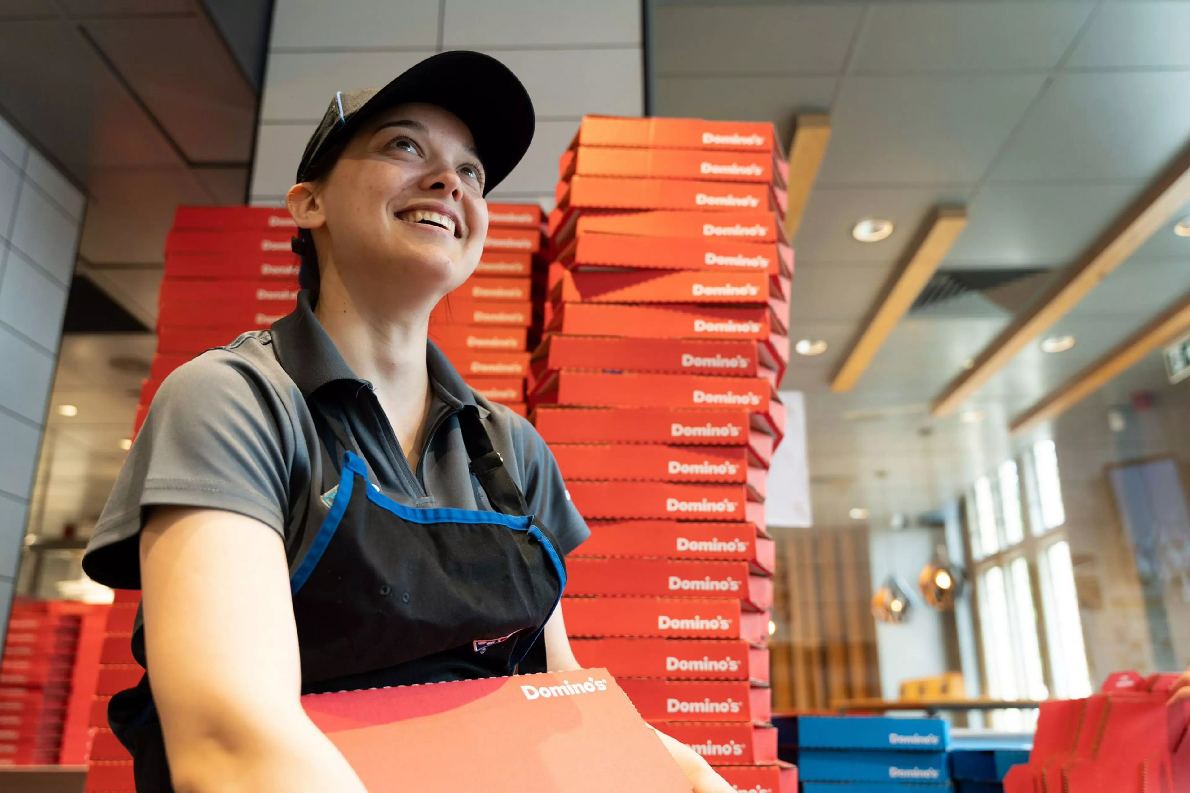 A dominos worker is in store carrying a pizza box, she is smiling and standing in front of a large stack of other pizza boxes.