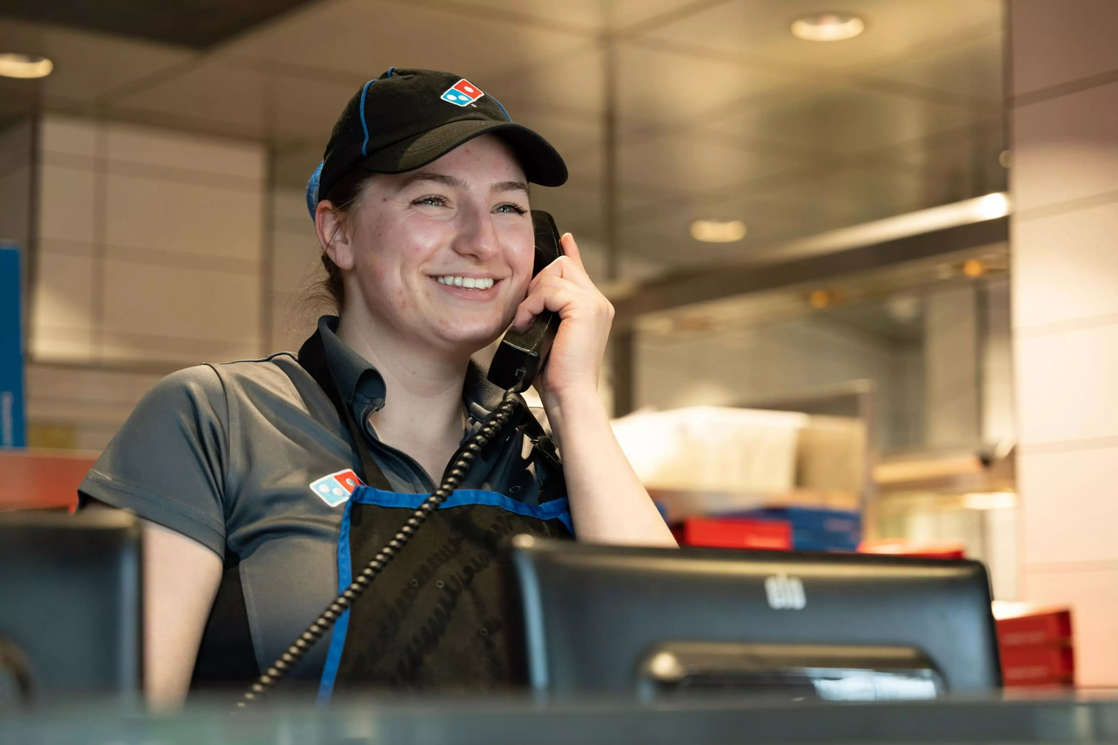 A woman is smiling while on the phone inside of a Domino's store.