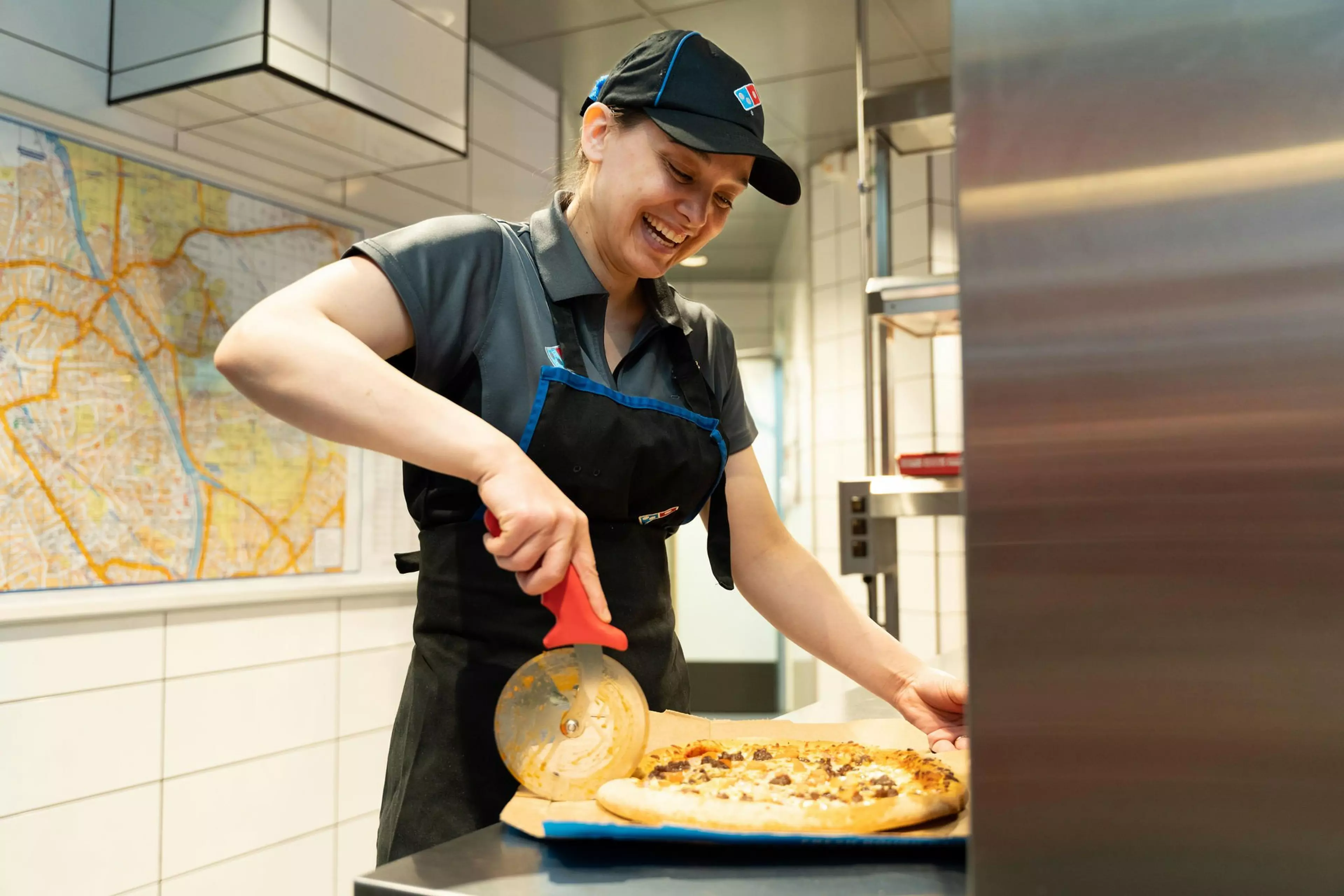 A Domino's worker is cutting a pizza with a pizza cutter in a Domino's store.