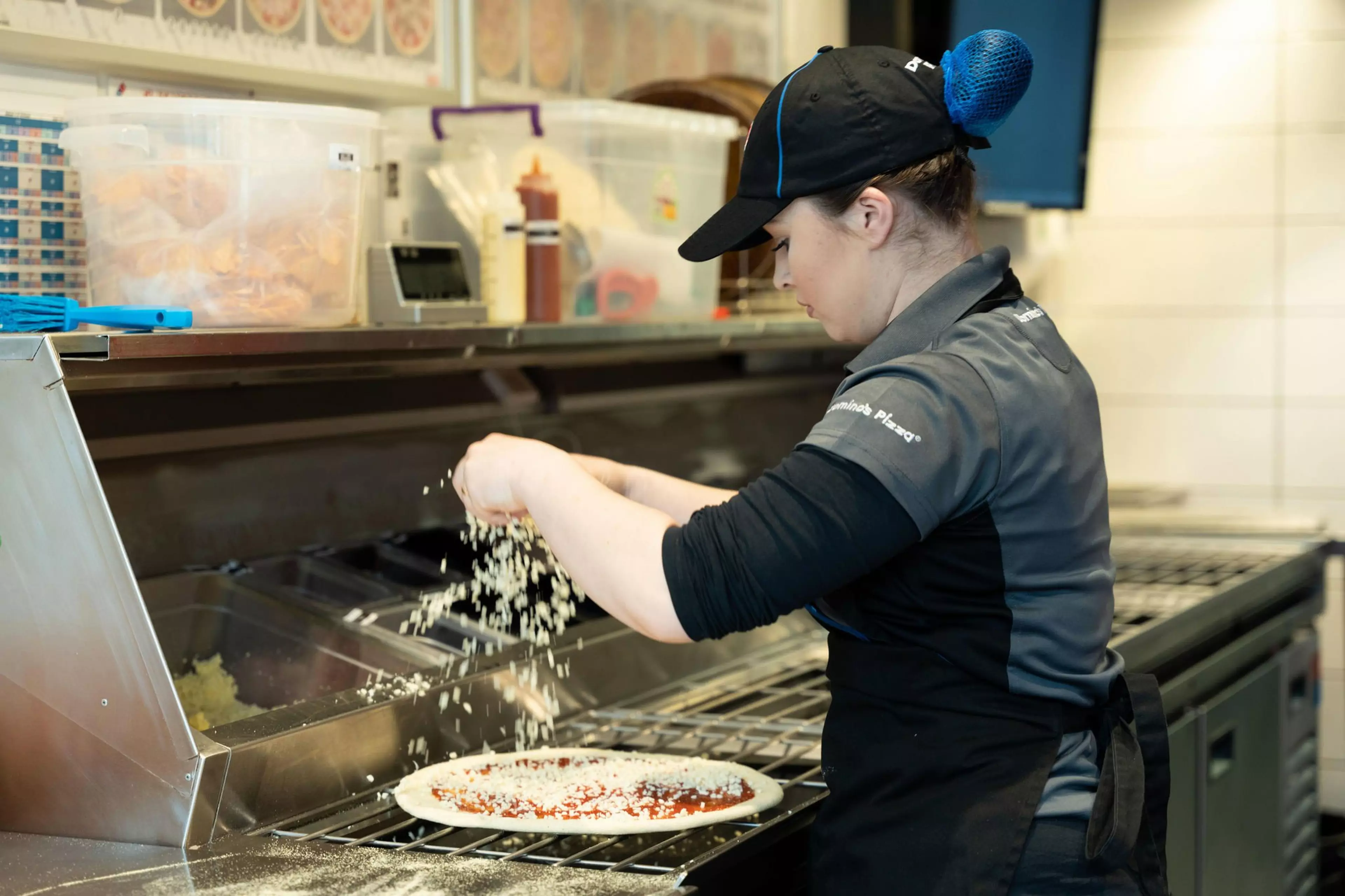 A Domino's worker is pictured in store sprinkling cheese on a pizza base.