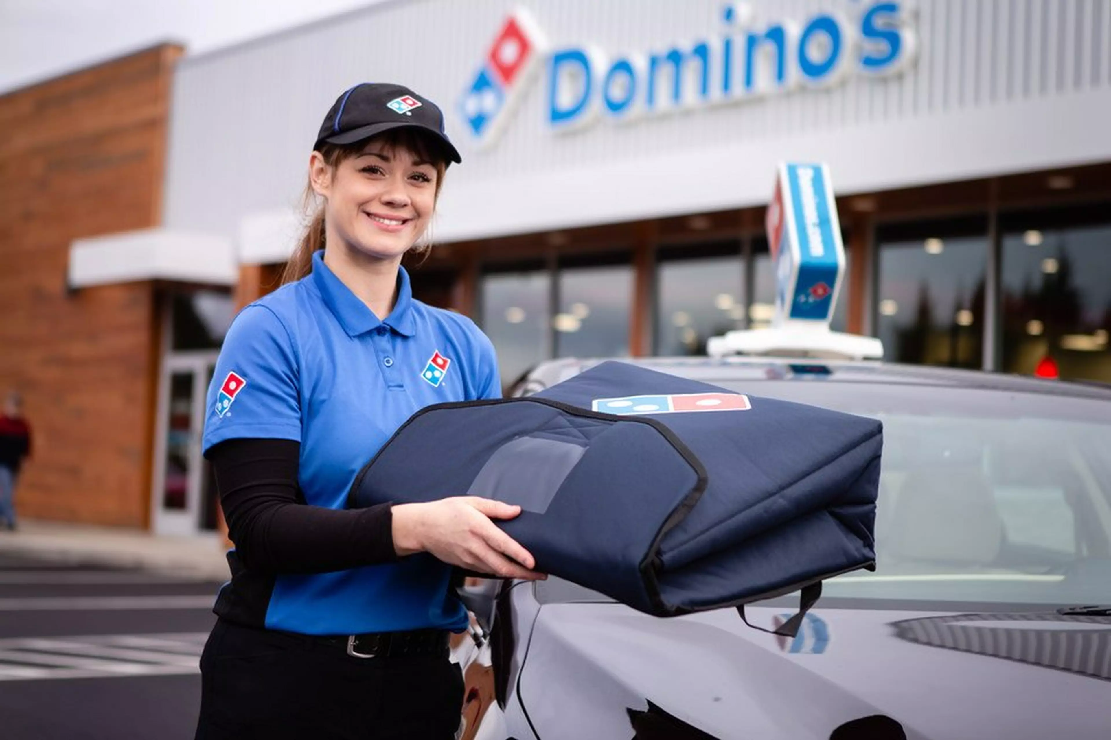 A smiling woman wearing a Domino's uniform is standing in front of a car holding a pizza delivery bag.