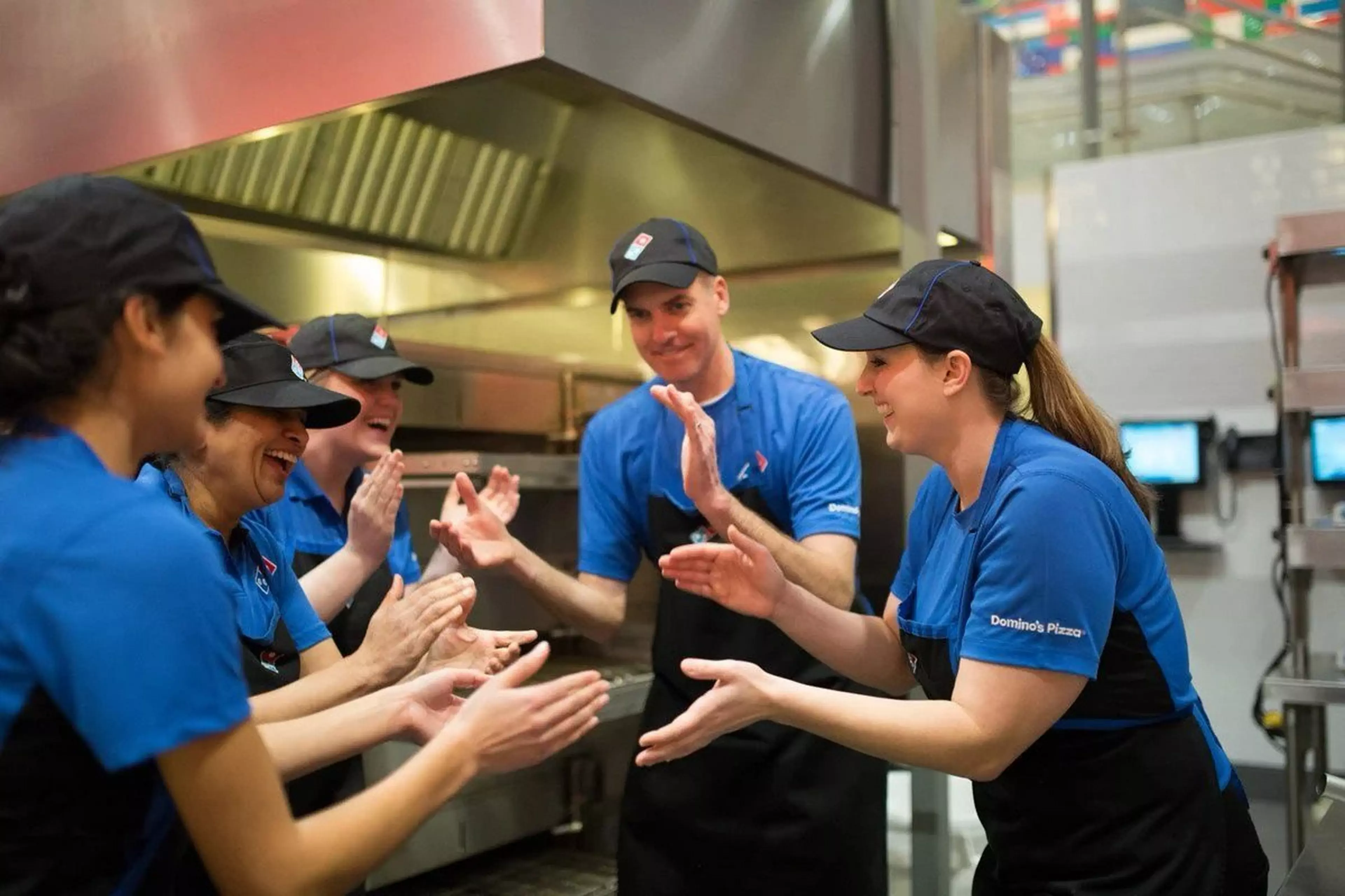 Five Domino's staff members standing in a circle in a Domino's store, they are all clapping.