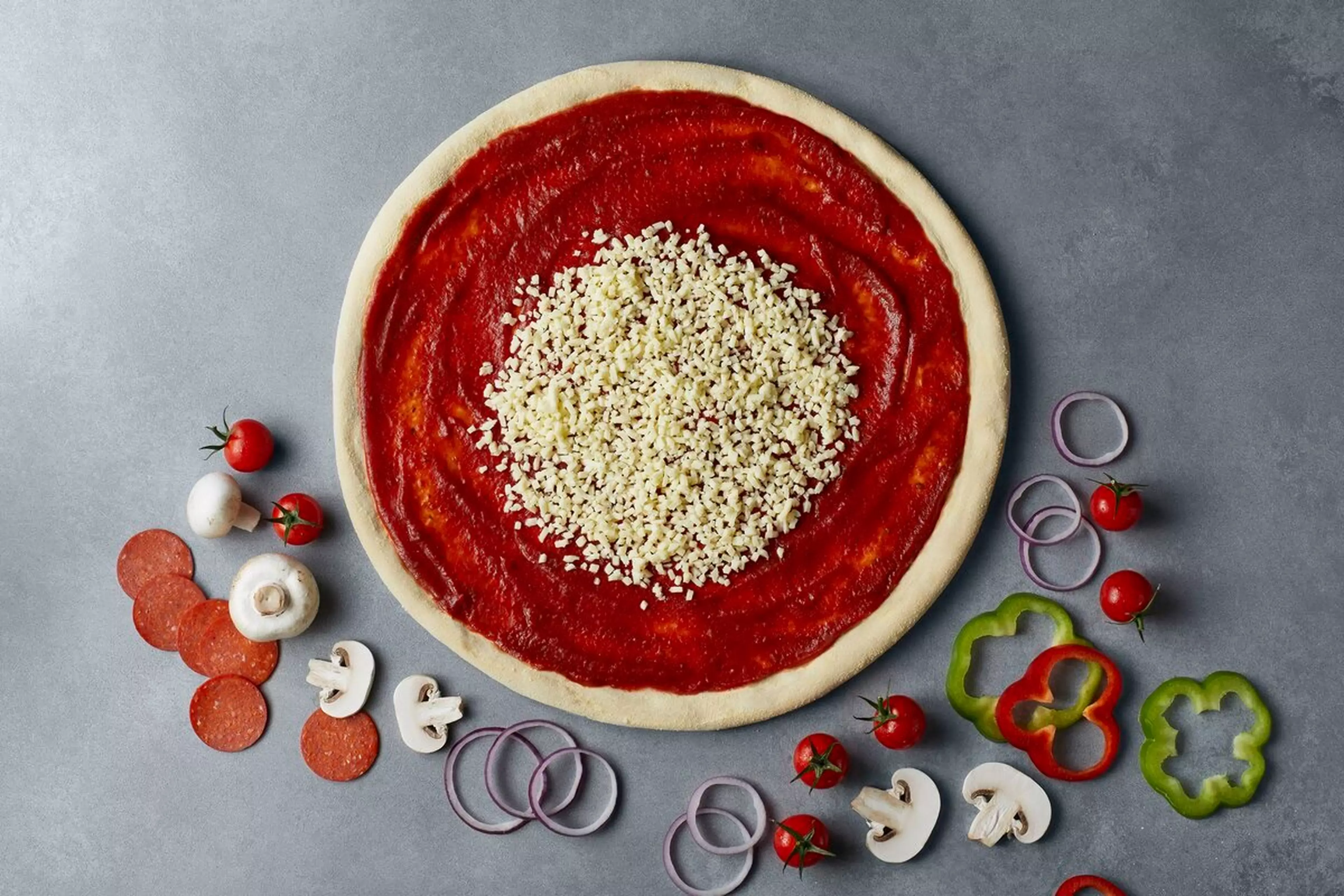 A pizza is in the process of being made, it has sauce and cheese on a base while there are various other topping arranged around it.